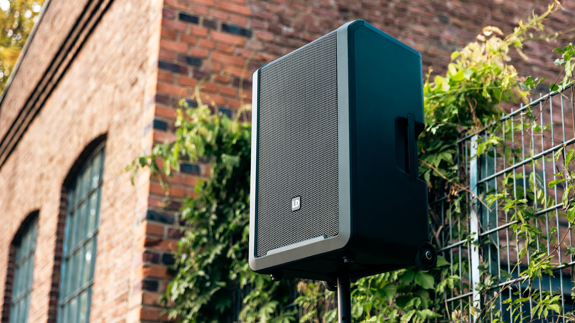 Enceinte sono portable LD Systems ANNY 12 sur pied en extérieur, fond de mur en briques et végétation