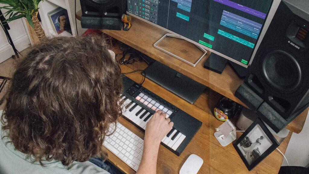 Producteur en home studio jouant sur un Novation Launchkey Mini 37 MK4 posé sur un bureau en bois, face à un écran affichant une session DAW, avec des enceintes de monitoring Adam Audio de part et d'autre.