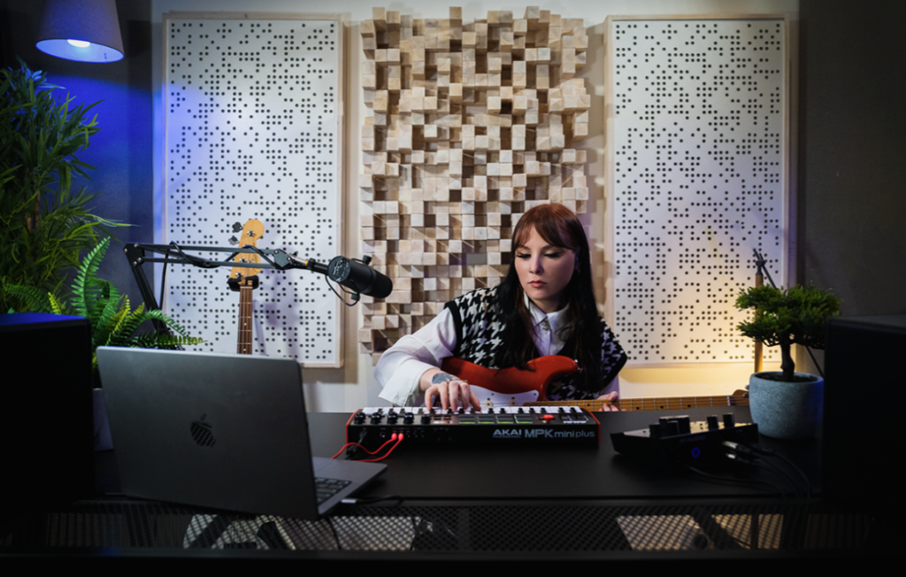 Musicienne jouant sur un Akai MPK Mini Plus dans un studio traité acoustiquement avec panneaux diffuseurs en bois, accompagnée d'une guitare électrique, d'un micro sur bras articulé et d'un ordinateur portable.