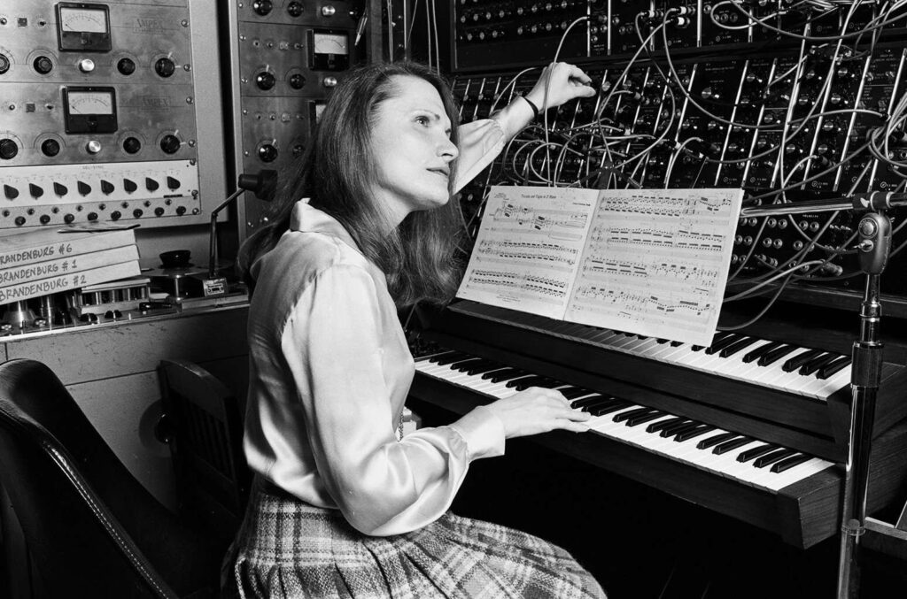 Wendy Carlos devant un synthétiseur modulaire Moog en studio, manipulant un câble de patch au-dessus d'un clavier et d'une partition, entourée de modules analogiques vintage