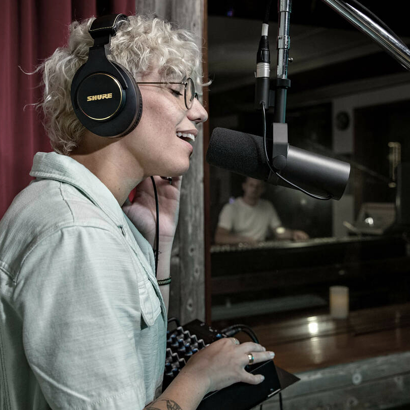 Micro dynamique Shure SM7B avec bonnette mousse, monté sur pied, utilisé par une animatrice portant un casque fermé dans un studio d’enregistrement